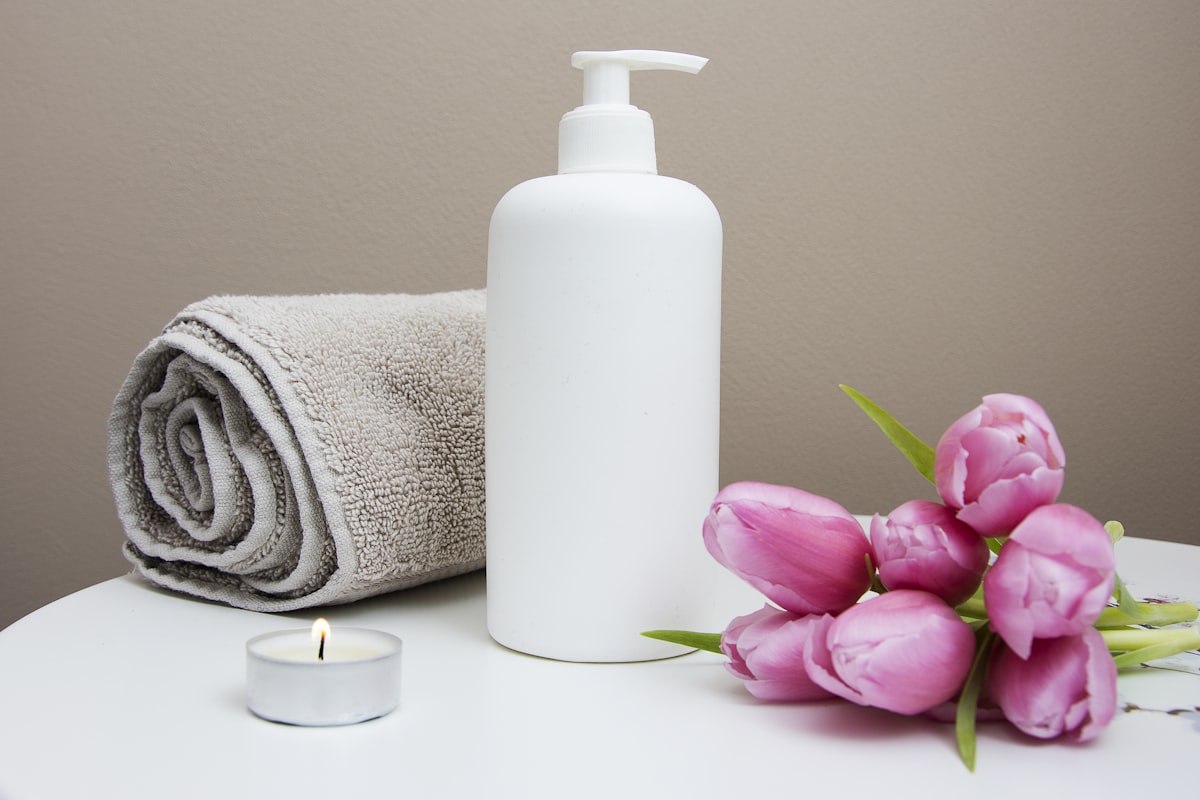 Lotion bottle with rolled towels and flowers on a neutral surface